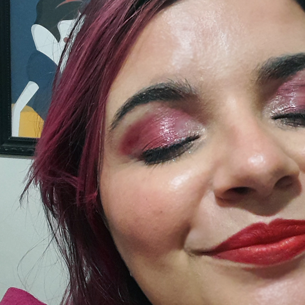 Close do rosto de uma mulher com maquiagem em tons de rosa cintilante nos olhos e batom vermelho intenso, usando delineado preto; ela aparece com os olhos fechados, destacando o brilho da sombra e a pele iluminada, com cabelo em tom rosado ao fundo.