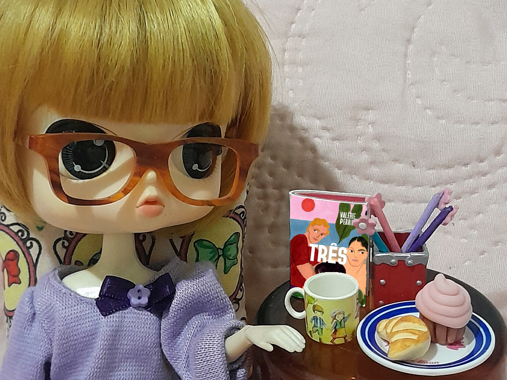 Boneca com óculos e roupa lilás ao lado de uma mesinha com café da manhã em miniatura, incluindo caneca, croissant e cupcake rosa, com o livro Três apoiado próximo aos objetos.