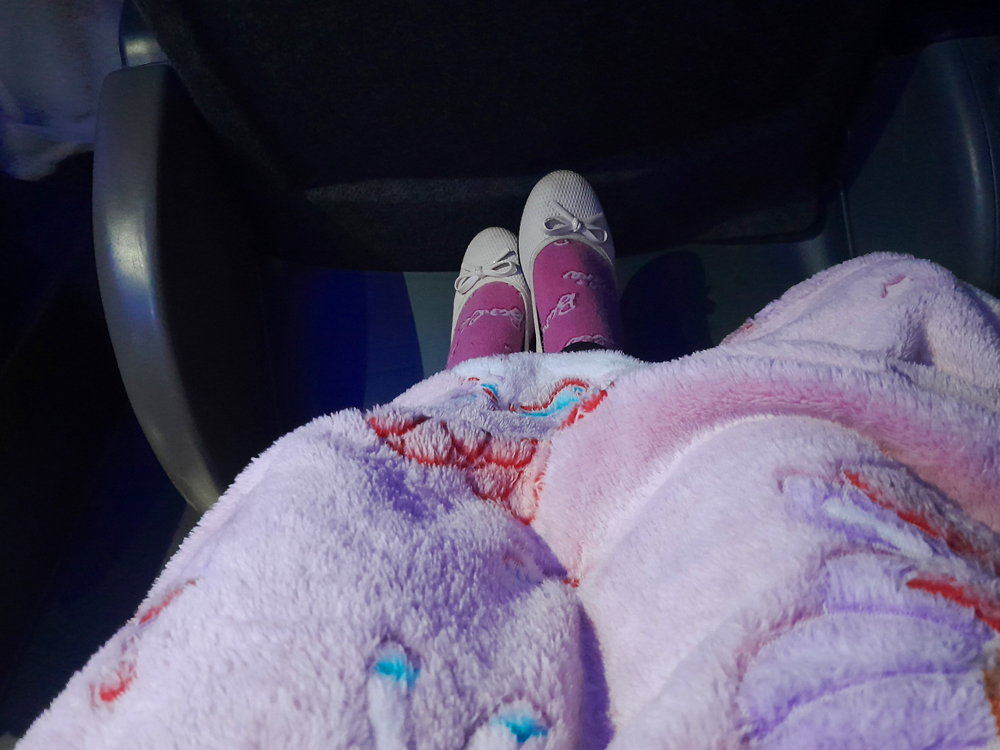 Visão de cima das pernas de uma pessoa sentada, usando meias rosas e sapatilhas claras com laço. Um cobertor macio em tons de rosa e lilás cobre o colo, criando uma cena aconchegante dentro de um ambiente interno.