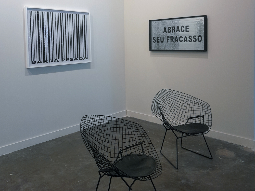 Canto de um estande da SP-Arte com duas cadeiras de metal preto em frente a obras de parede. Uma das obras traz um código de barras estilizado e outra, em fundo cinza, exibe a frase ABRACE SEU FRACASSO em destaque.