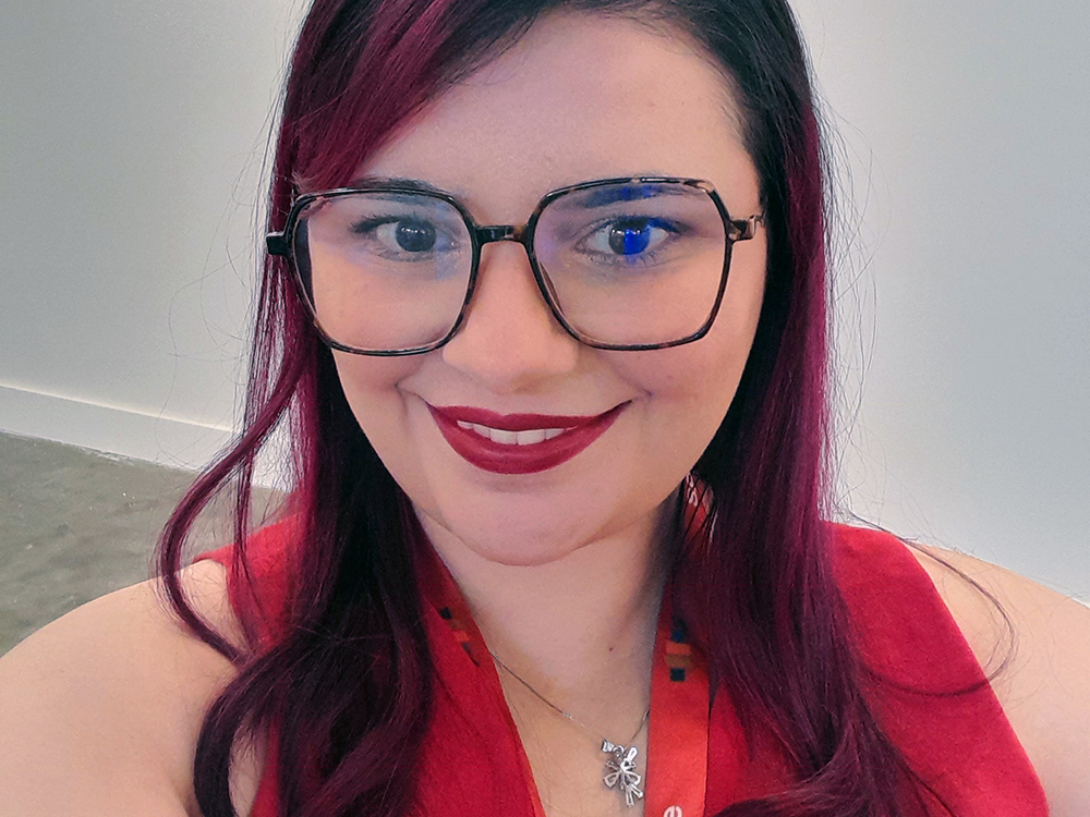 Selfie de uma mulher sorrindo em ambiente interno de galeria. Ela tem cabelos pretos e rosas, usa óculos de armação grande, batom vermelho e blusa sem mangas também vermelha, com crachá da SP-Arte no pescoço. Ao fundo, parede branca e piso de concreto.