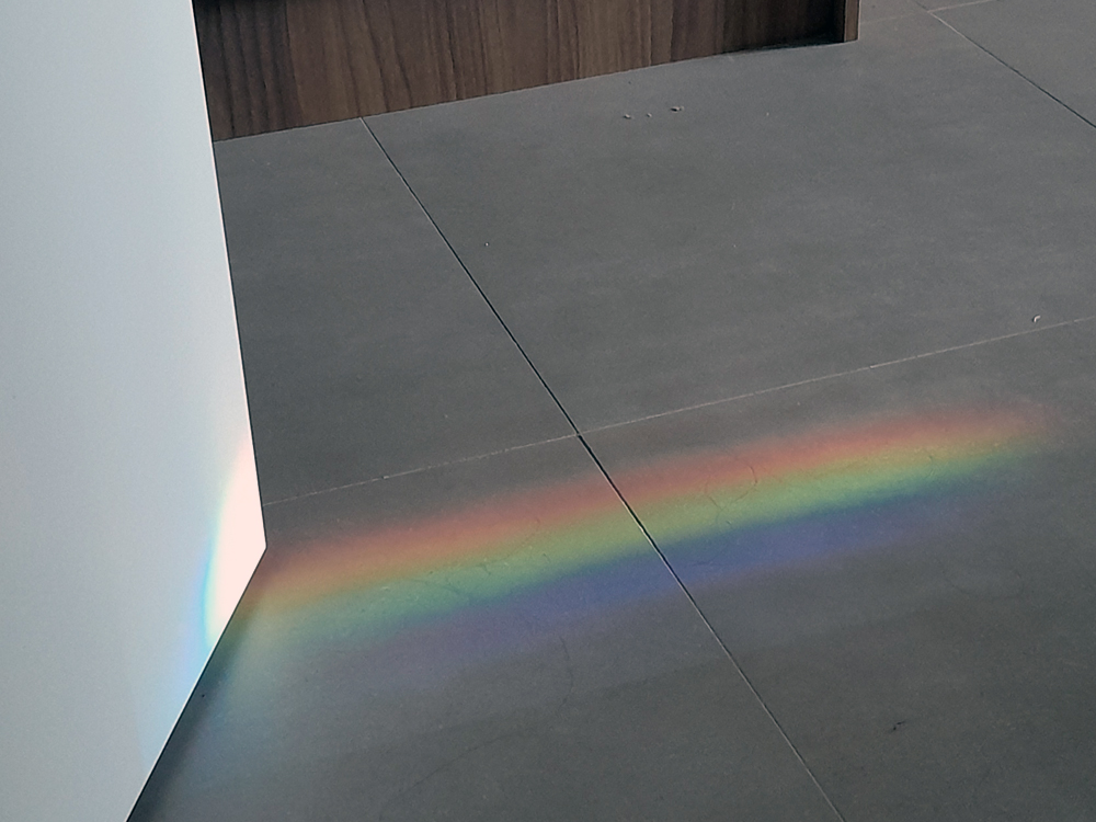Faixa de luz formando um arco-íris projetado no chão de concreto ao lado de uma parede branca, criando um efeito colorido e delicado em contraste com o ambiente neutro.