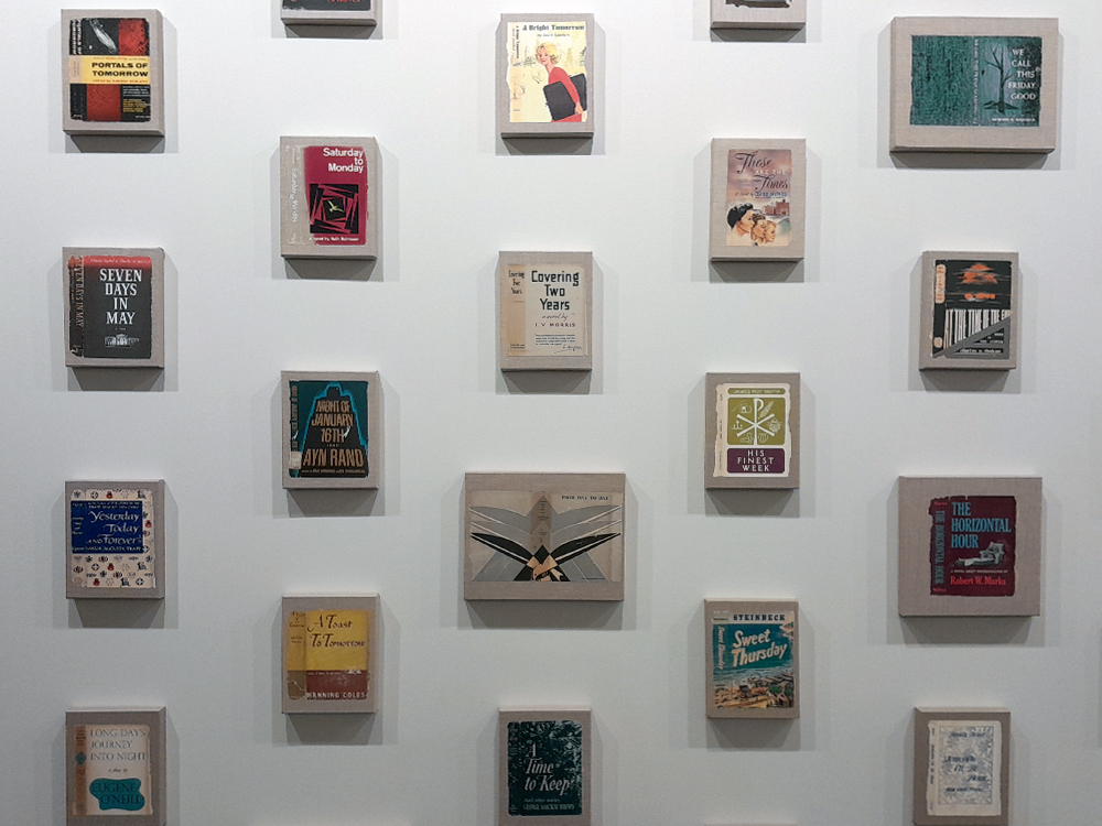 Parede de galeria com uma grade organizada de pequenas obras emolduradas, cada uma contendo capas de livros ou composições gráficas. As peças estão dispostas em linhas simétricas sobre fundo branco, criando um efeito visual de coleção.