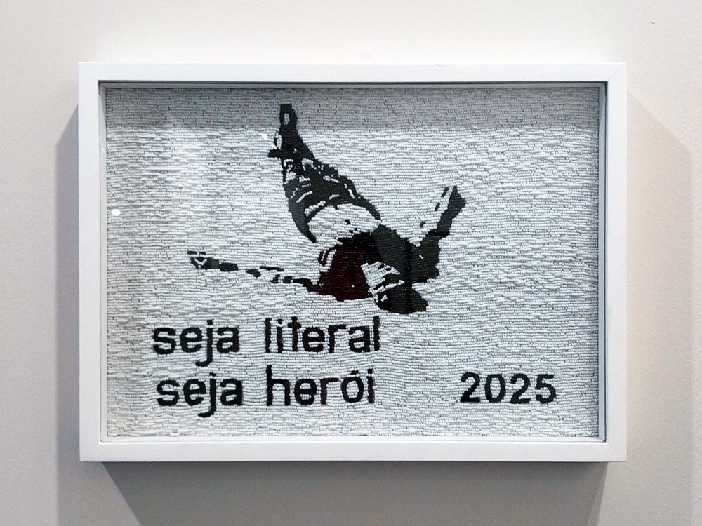 Obra de Marta Neves emoldurada com fundo claro e textura de miçangas, seu material, mostrando a silhueta de uma figura em queda. Abaixo da imagem, lê-se a frase seja literal seja herói 2025 em letras pretas.