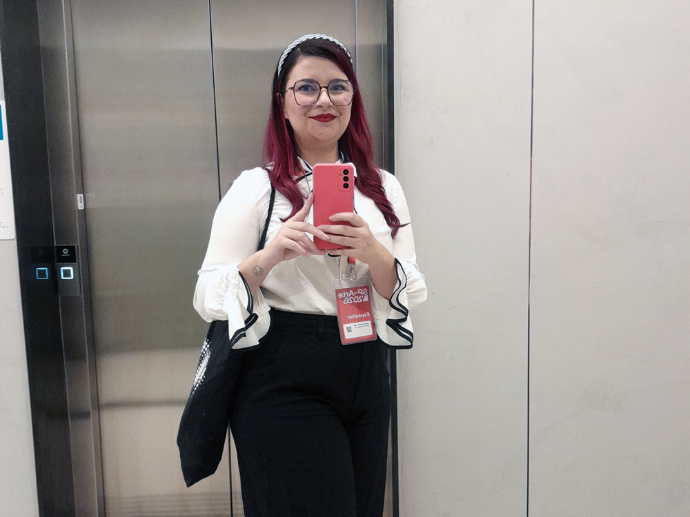 Foto no espelho de elevador: uma mulher posa sorrindo, usando blusa branca com mangas bufantes e gola de laço, calça preta de cintura alta e bolsa preta no ombro. Ela segura um celular rosa e usa um crachá da SP-Arte pendurado no pescoço.