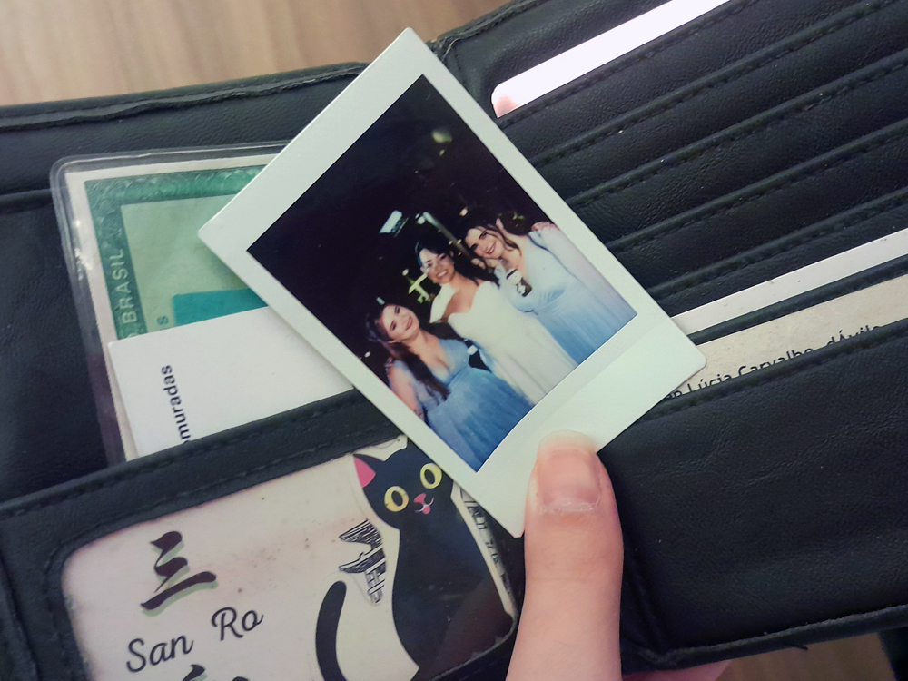 Carteira aberta mostrando documentos e cartões, enquanto uma mão segura uma foto instantânea (tipo polaroid) de três mulheres sorrindo juntas. A carteira tem estampa com um gatinho preto e detalhes pessoais visíveis.