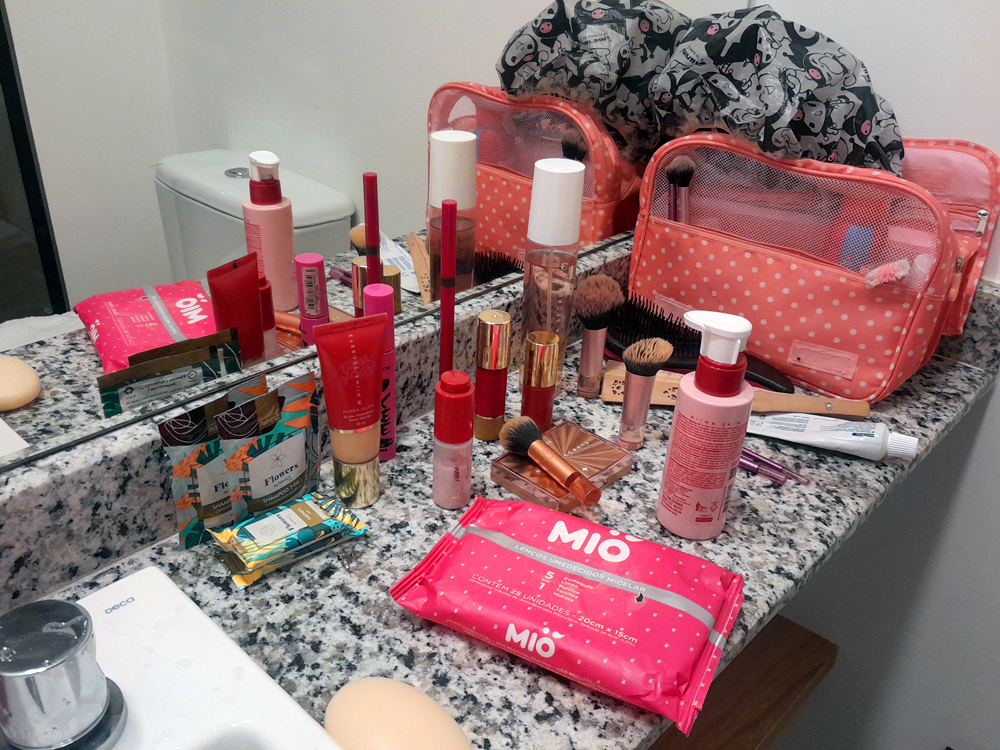 Bancada de banheiro com diversos itens de maquiagem e cuidados pessoais espalhados: pincéis, batons, base, lenços umedecidos e frascos de skincare. Ao fundo, um nécessaire rosa com bolinhas e um espelho refletem os objetos, criando sensação de rotina de beleza.