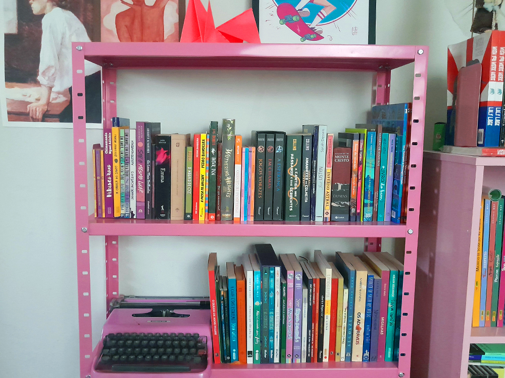 Estante de livros rosa com duas prateleiras cheia de livros coloridos, uma máquina de escrever antiga também rosa na parte inferior, ao lado de uma parede decorada com obras de arte em papel.