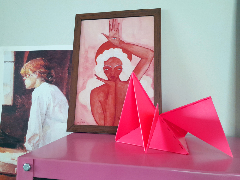 Imagem com três coisas diferentes sobre uma superfície cor de rosa metálica, Um objeto decorativo em acrílico simulando dobraduras na cor rosa neon, feitos de formas geométricas triangulares, em frente a duas obras de arte: uma ilustração de mulher com pele avermelhada e cabelo branco, com um olho desenhado na palma da mão erguida, e outra pintura de uma figura de perfil próxima a uma janela.