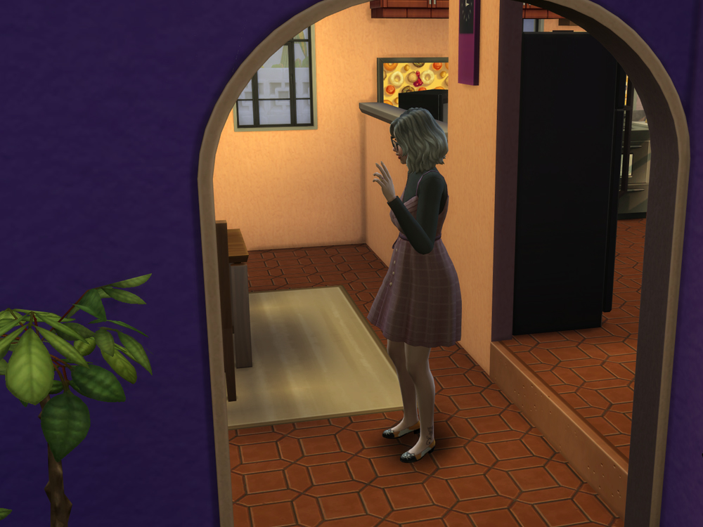 Personagem de estilo The Sims aparece em pé dentro de uma casa, vista através de um arco na parede roxa. Ela tem cabelo curto ondulado, usa óculos, blusa escura de manga longa e saia xadrez em tons rosados, com sapatilhas. O ambiente tem piso de cerâmica avermelhada, paredes em tom pêssego e, ao fundo, uma cozinha com geladeira preta e quadro decorativo na parede. A personagem parece concentrada, com uma das mãos levantadas como se estivesse pensando ou interagindo com algo no jogo.