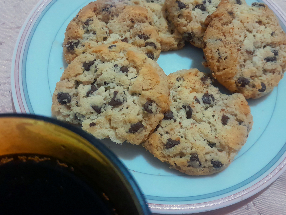 Prato com biscoitos caseiros com gotas de chocolate, levemente dourados, acompanhados por um copo de Coca Cola ao lado, parcialmente visível na lateral da imagem.