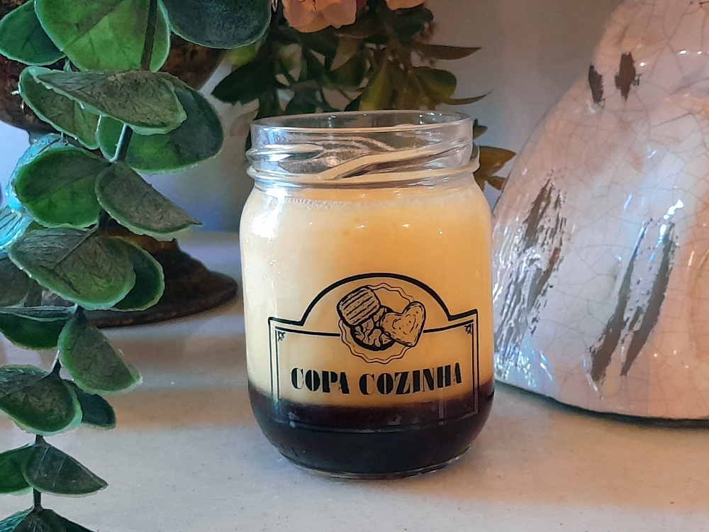 Pote de vidro com rótulo Copa Cozinha contendo um pudim de leite caramelizado, cercada por plantas e objetos decorativos de estética antiga.