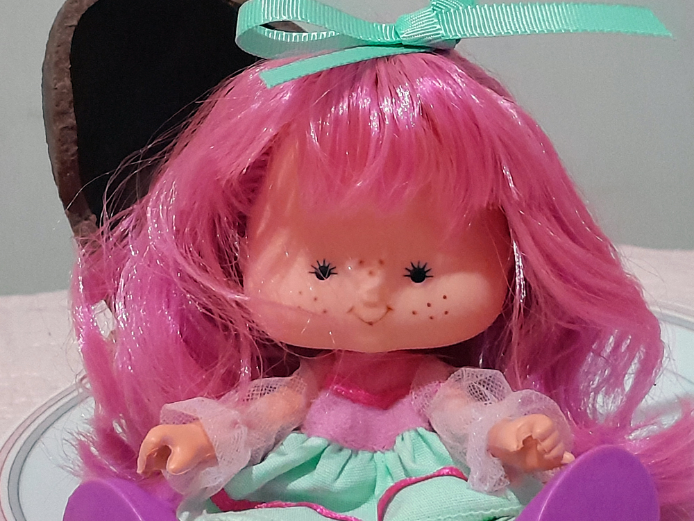 Boneca com cabelo rosa vibrante e laço verde, usando vestido delicado em tons pastel, com expressão sorridente e bochechas com sardinhas, com um ovo de páscoa de chocolate visto parcialmente atrás.