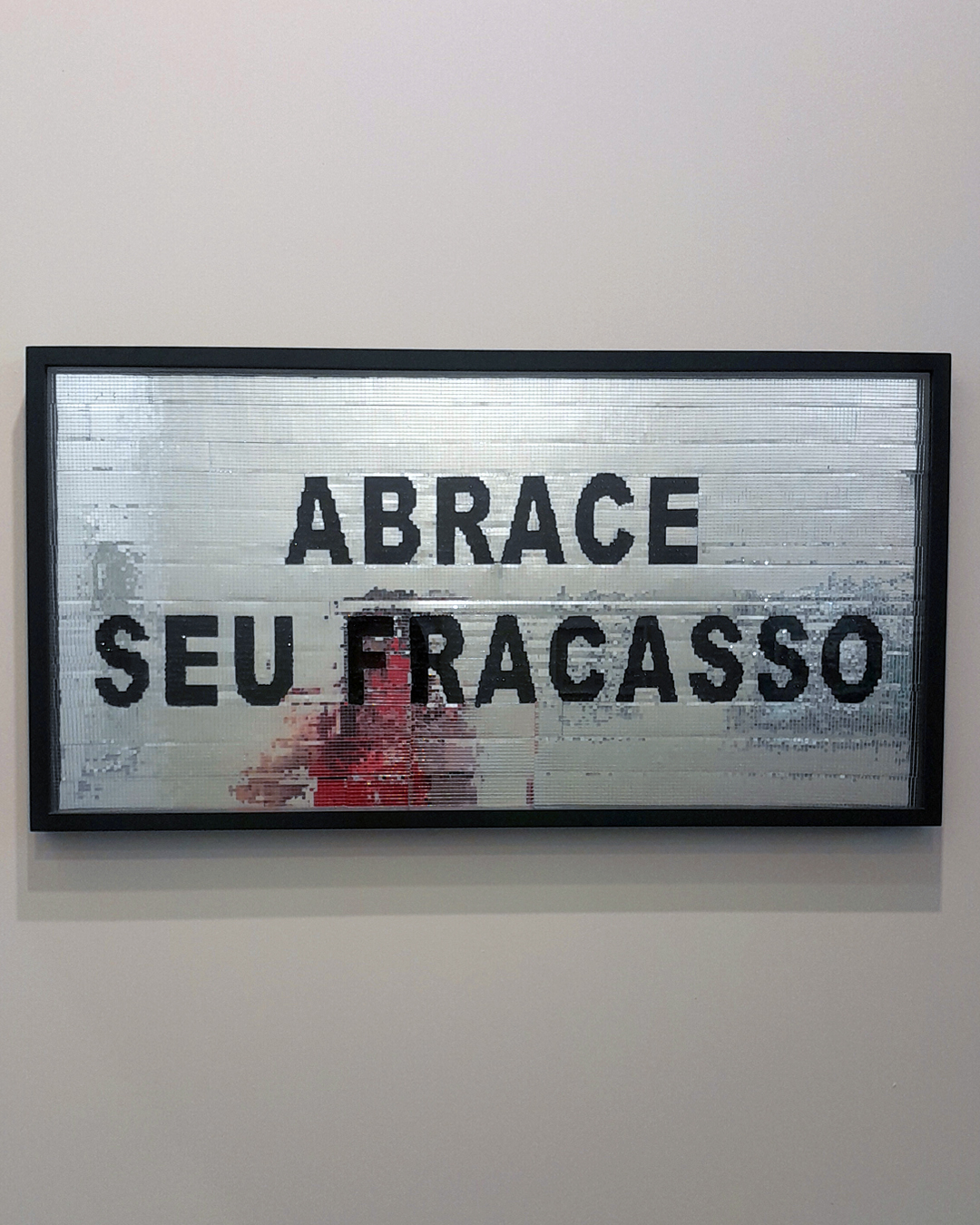 Quadro com a frase 'Abrace seu fracasso' em letras pretas em paetês, com fundo de espelhos refletindo a pessoa que o fotografa, distorcida, e o fundo de um estande da SP-Arte.