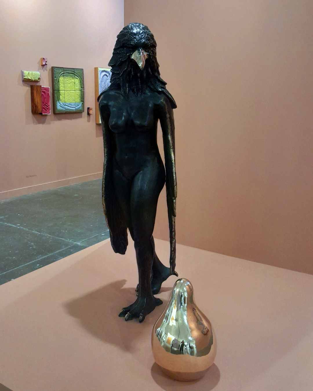 Escultua de uma figura feminina com cabeça de pássaro, escura, com peça dourada em formato de pera em sua frente, em um estande cor de pêssego da SP-Arte.