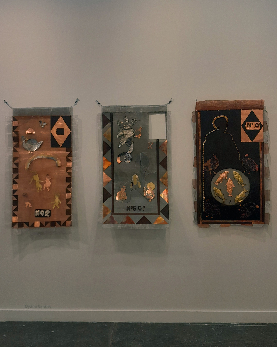 Três obras de arte de Dyana Santos expostas na parede, com detalhes em metal e cores quentes, parecendo visualmente estandartes contemporâneos.