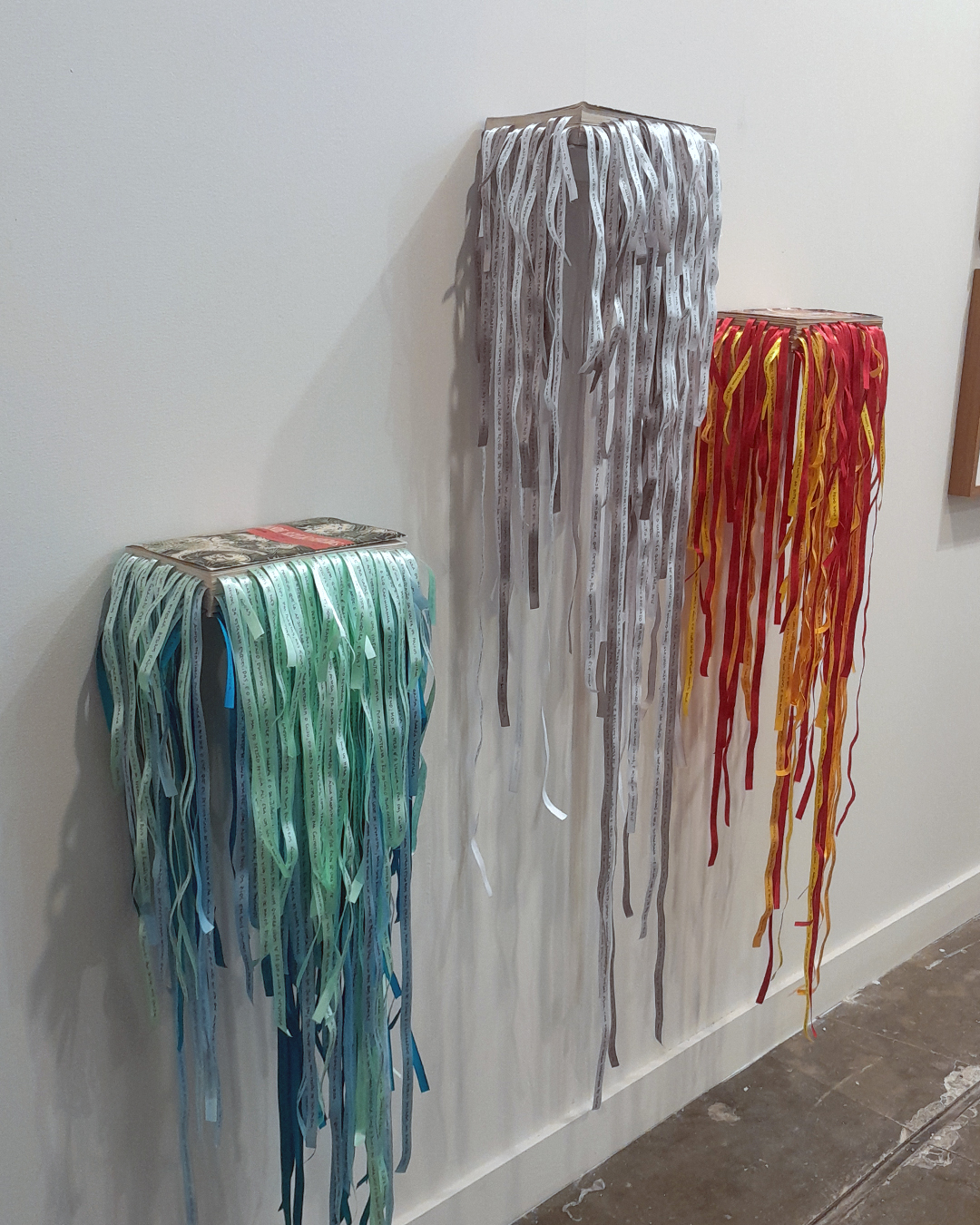 Trabalho de Marilá Dardot composto por livros fixados na horizontal na parede, dos quais saem fios brilhantes nas cores verde, prata e alaranjado.