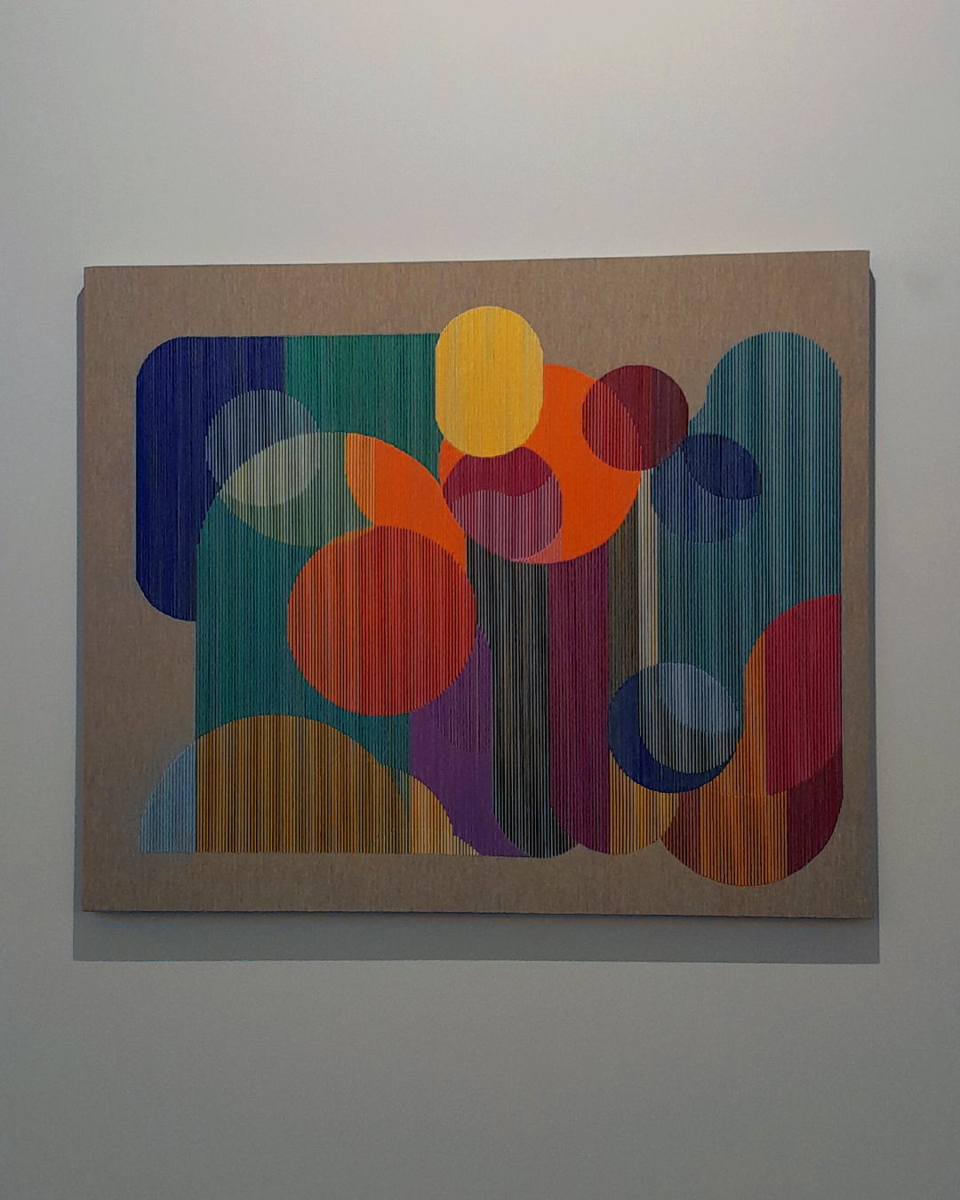 Imagem de quadro abstrato com formas geométricas coloridas e formadas por linhas finas em diferentes cores, bordadas em fundo bege.