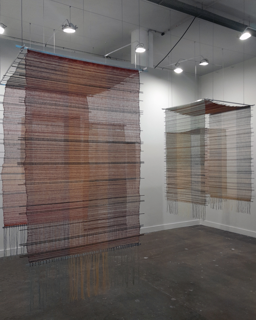 Instalações de arte de Maria Appleton com peças de tecido suspensas em ambiente interno, destacando o uso de cores neutras e o conceito de espaço e textura.