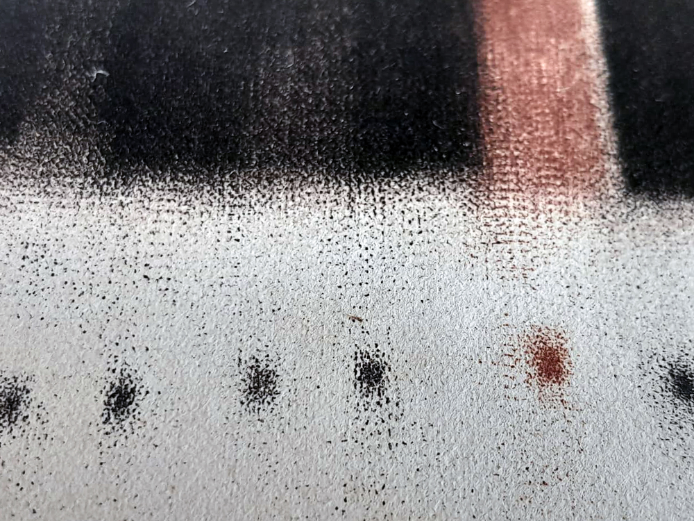 Close em uma superfície branca texturizada com pequenos pontos pretos alinhados em sequência, abaixo de faixas verticais escuras e rosadas. A fotografia abstrata destaca padrões, granulação e linhas suaves formadas pelas marcas na imagem.