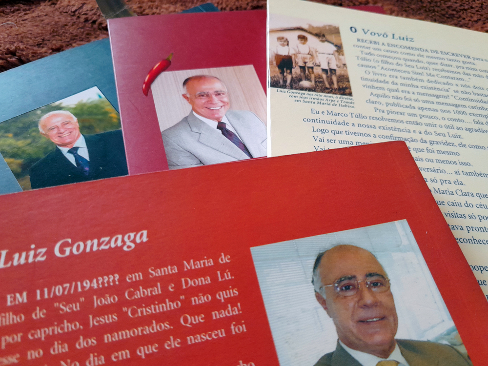 Close nas quarta capas de exemplares das coletâneas de causos de Luiz Gonzaga Viana Lage espalhados sobre uma superfície marrom felpuda. As capas em tons de vermelho e bege trazem fotografias do autor sorrindo, além de trechos de texto e imagens antigas de família.