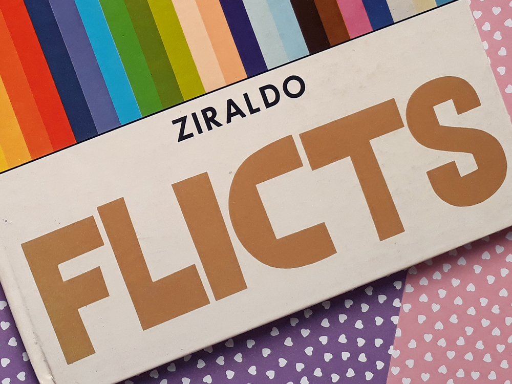 Close na capa do livro com enquadramento destacando título em letras grandes e parte das faixas coloridas que ocupam o topo da capa. Ao fundo, aparecem papéis decorados em tons de rosa e lilás com pequenas estampas de corações.
