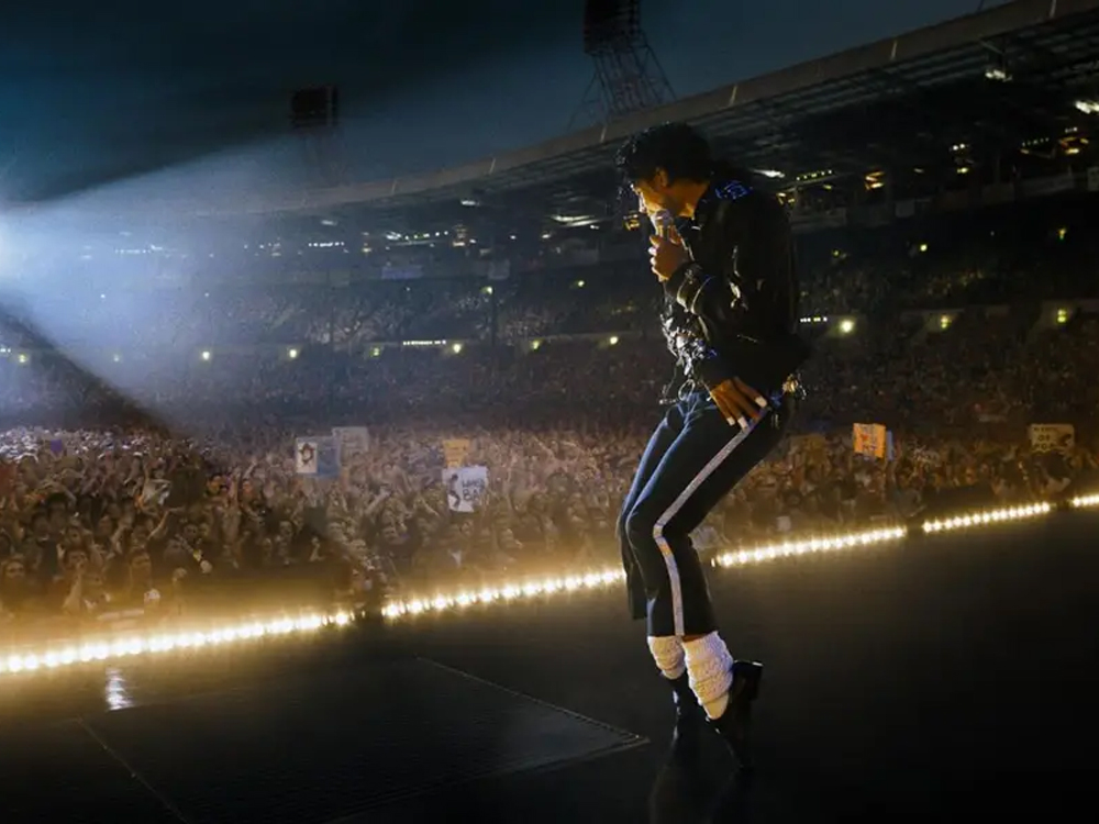 Cena de show: artista caracterizado como Michael Jackson dança no palco diante de uma multidão iluminada. Ele usa figurino preto com detalhes brilhantes, meias claras e sapatos escuros, enquanto refletores e luzes do estádio destacam a performance.