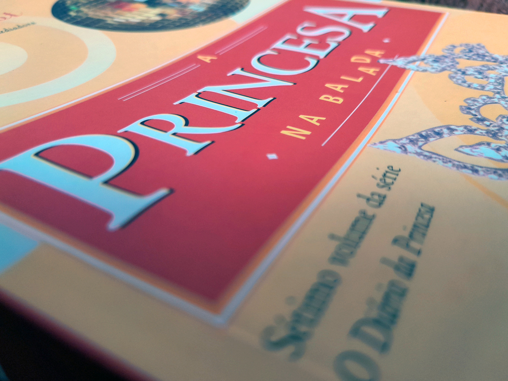 Close da capa do livro A Princesa na Balada, de Meg Cabot, fotografada em ângulo baixo. O destaque é a faixa central em tom laranja-avermelhado com o título da obra, enquanto o restante da capa aparece suavemente desfocado ao fundo.