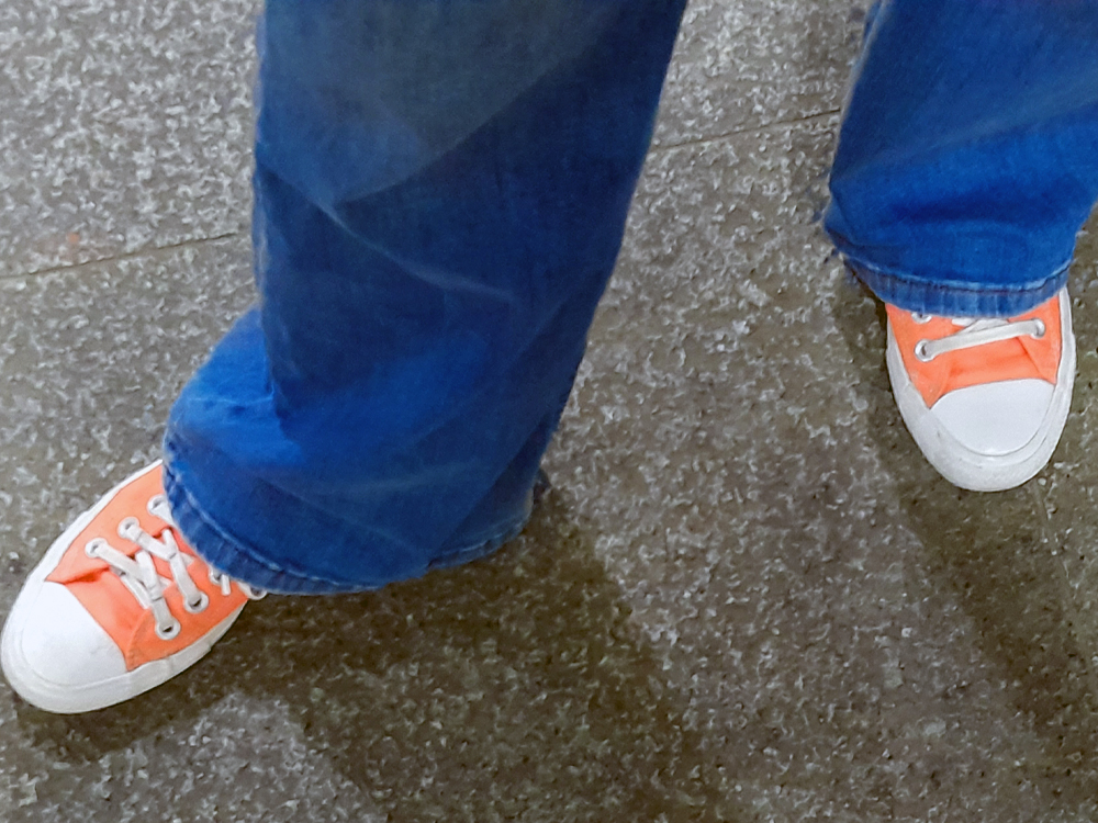 Vista de cima dos pés de uma pessoa usando tênis laranja-neon com ponteiras brancas e calça jeans azul, em contraste com o piso cinza mesclado.