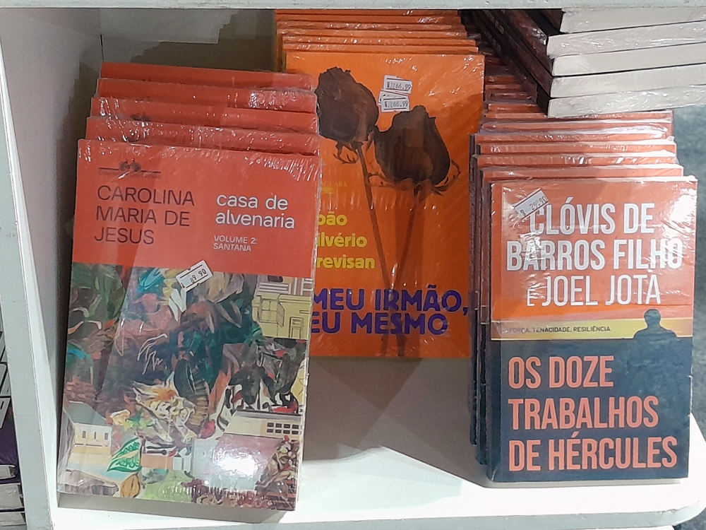 Prateleira de livraria com diversos livros de capas predominantemente laranjas. Em destaque estão exemplares de Casa de Alvenaria, de Carolina Maria de Jesus, além de outros títulos organizados lado a lado, formando uma composição marcada pelos tons quentes.