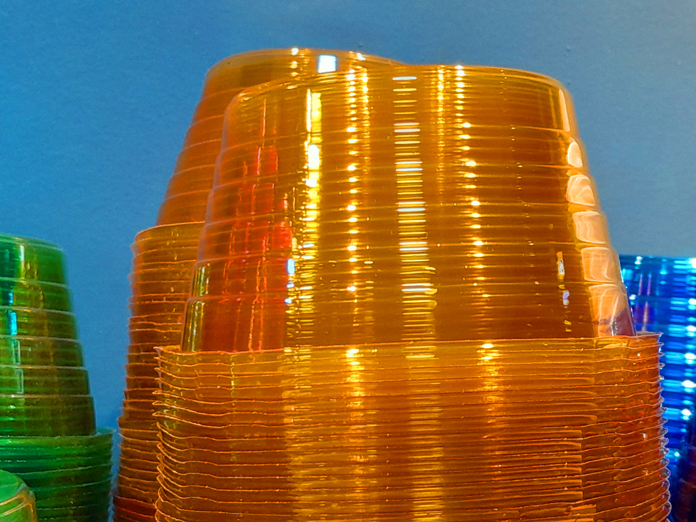 Pilhas de copinhos para sorvete, um sobre o outro encaixados com a boca virada pra baixo, sendo duas dela em cor de laranja neon en destaque e um pequeno vislumbre de uma pilha verde e uma azul nas laterais.