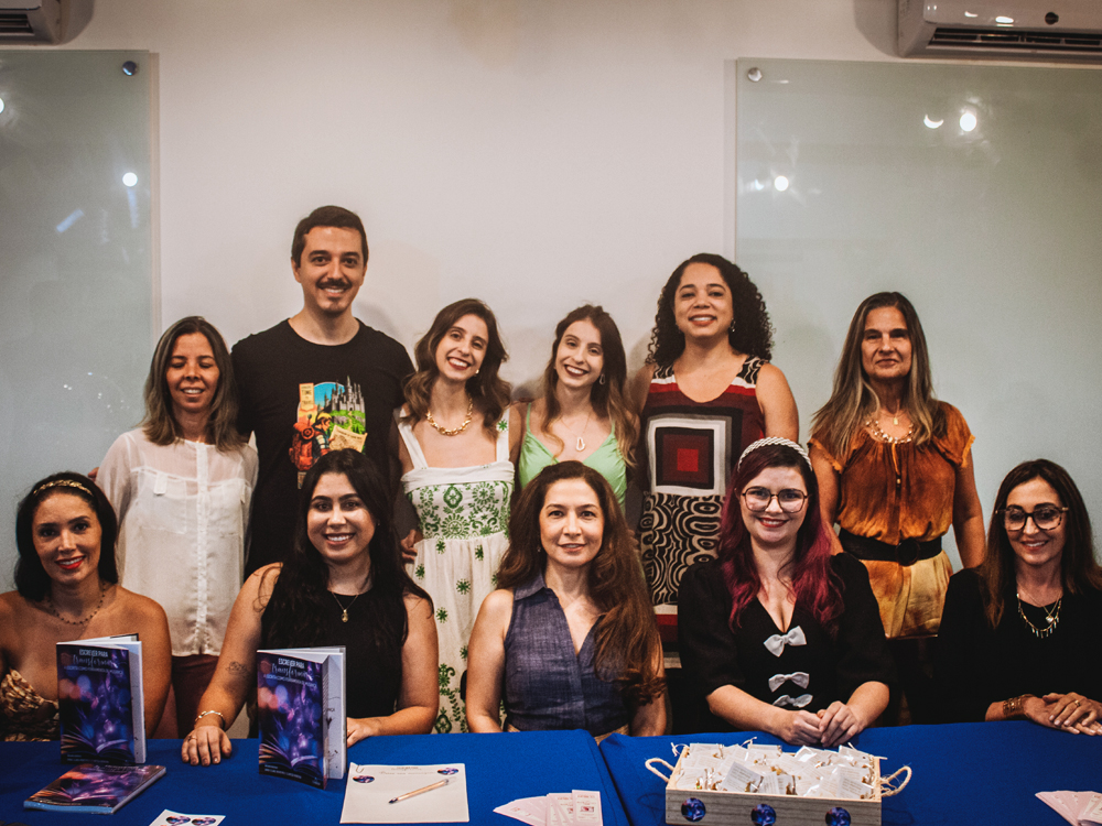 Fotografia de grupo durante o lançamento do livro Escrever para Transformar: A Escrita como Ferramenta de Mudança. Seis pessoas estão sentadas atrás de uma mesa coberta por toalha azul, enquanto outras seis posam em pé ao fundo. Exemplares do livro, materiais de divulgação e brindes estão distribuídos sobre a mesa. Todos sorriem para a câmera em um ambiente de evento literário.