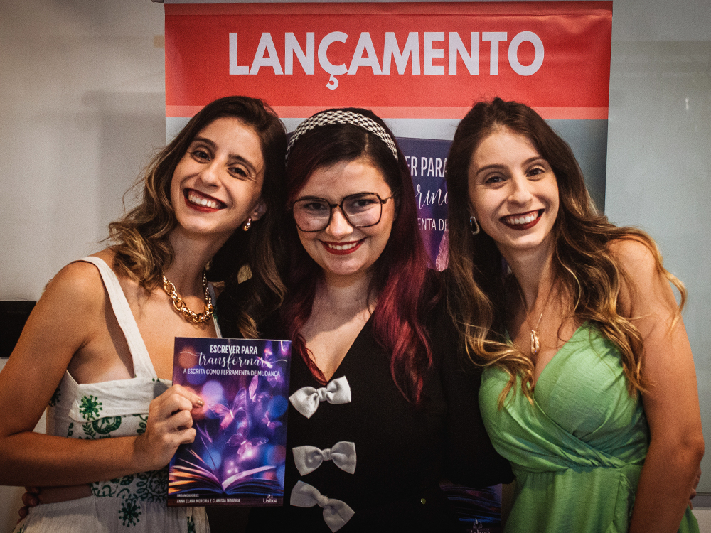 Três mulheres posam sorrindo diante de um banner com a palavra Lançamento. A mulher ao centro, de cabelos cor-de-rosa, segura um exemplar do livro Escrever para Transformar: A Escrita como Ferramenta de Mudança, enquanto as outras duas, gêmeas idênticas, a abraçam pelos ombros.