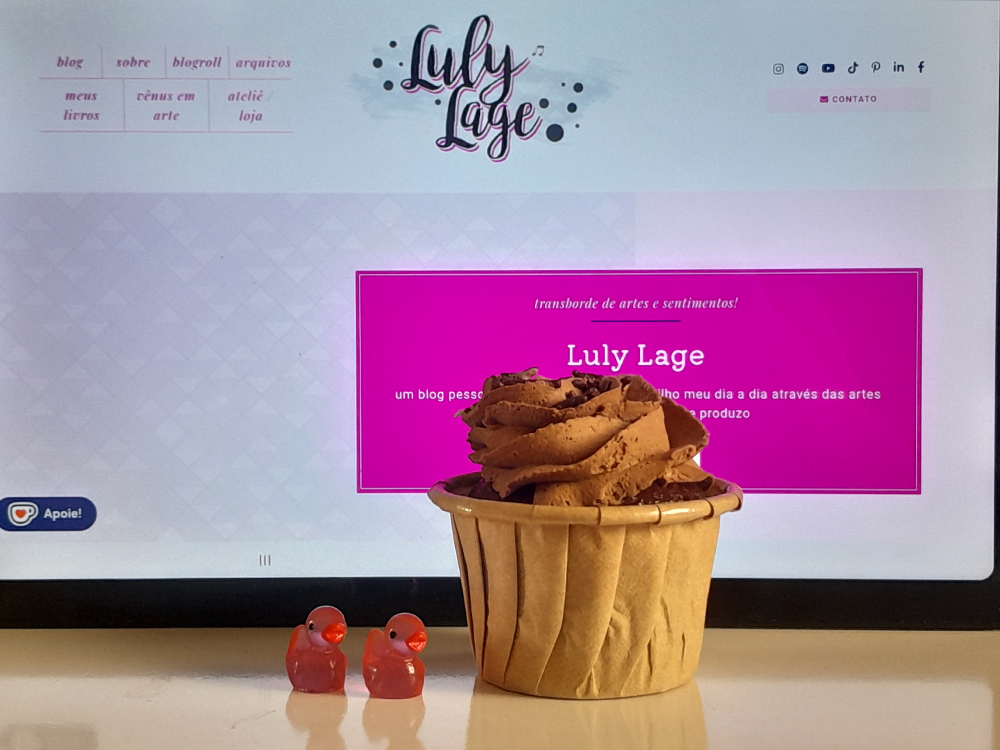 Um cupcake de chocolate com cobertura em espiral está sobre uma mesa branca, posicionado diante de um monitor que exibe a página inicial do blog Luly Lage. Diferentemente da foto anterior, o cupcake aparece sem velas acesas. Dois pequenos patinhos de borracha cor-de-rosa representando o número 22 estão posicionados à frente do doce.