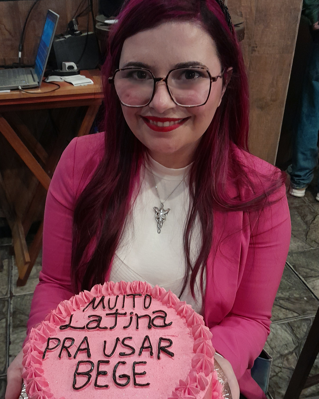 Luly Lage sorri em seu aniversário de trinta e seis anos segurando um bolo de aniversário rosa decorado com a frase Muito latina pra usar bege. Ela veste blazer rosa, blusa branca e um colar prateado, com os cabelos em tom rosa escuro e maquiagem em tons de vermelho e roxo.