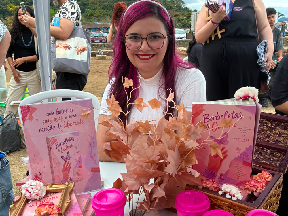 Luly Lage sentada em sua mesa de autógrafos da Casa Gueto durante a FLIP 2026, sorrindo para a câmera. Sobre a mesa há exemplares de Borboletas de Outono, displays do livro, marcadores de página, copos personalizados em tom de rosa, uma cesta decorativa e um arranjo de folhas secas que remete ao tema da obra. Ao fundo aparecem outros autores, visitantes e a paisagem externa de Paraty.