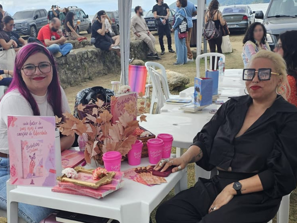 Luly Lage e Marianna Roman sentadas lado a lado durante uma sessão de autógrafos na FLIP 2026. As duas estão atrás de uma mesa branca decorada com materiais de divulgação, exemplares de livros, copos personalizados e um arranjo de folhas secas em tons de outono. Ao fundo, visitantes conversam e observam os estandes montados próximos à orla de Paraty.