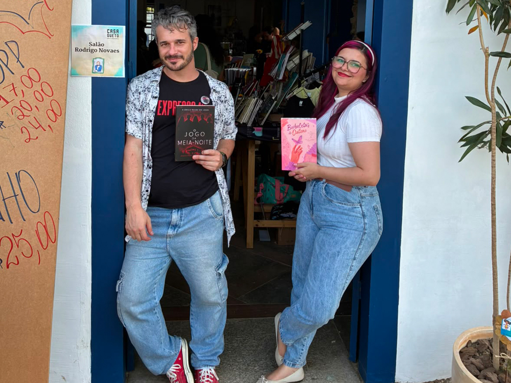 Luly Lage posa ao lado de Paulo Pera na entrada da Casa Gueto durante a FLIP 2026. Cada um segura um exemplar de seu próprio livro: Luly exibe Borboletas de Outono, enquanto ele apresenta Jogo Meia-Noite. Ambos sorriem para a câmera em frente à porta azul do espaço, onde também é possível ver a placa do Salão Rodrigo Novaes e parte do interior da livraria ao fundo.