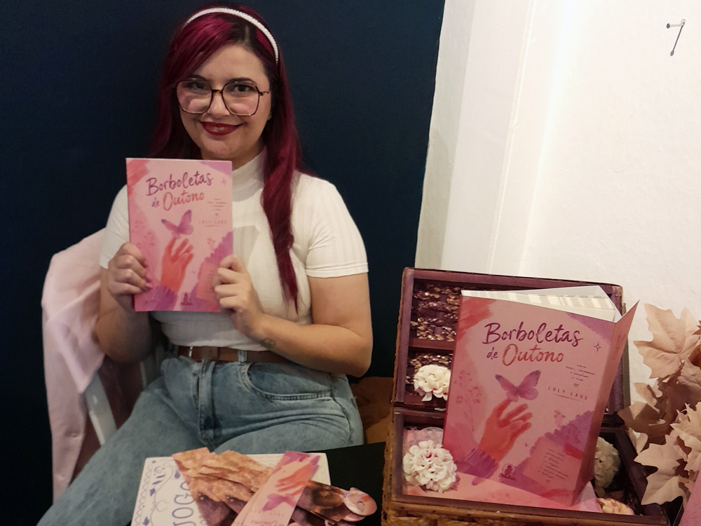 Luly sentada em sua mesa de autógrafos Escreva, Garota durante a FLIP 2026, segurando um exemplar de Borboletas de Outono. Ao lado dela há uma caixa decorativa de madeira com outro exemplar do livro em destaque, flores artificiais e materiais de divulgação. A autora sorri enquanto posa diante de um painel azul escuro, usando blusa branca, jeans claro, óculos de armação grande e cabelos rosa presos por uma tiara branca.