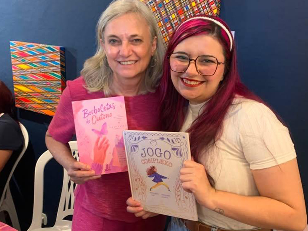Posando ao lado da escritora Katia Sentinaro durante a FLIP 2026. As duas sorriem para a câmera segurando exemplares de seus livros trocados: Katia exibe Borboletas de Outono, de capa rosa com uma borboleta em destaque, enquanto Luly segura Jogo Complexo. Ao fundo há uma parede azul decorada com obras de arte coloridas.