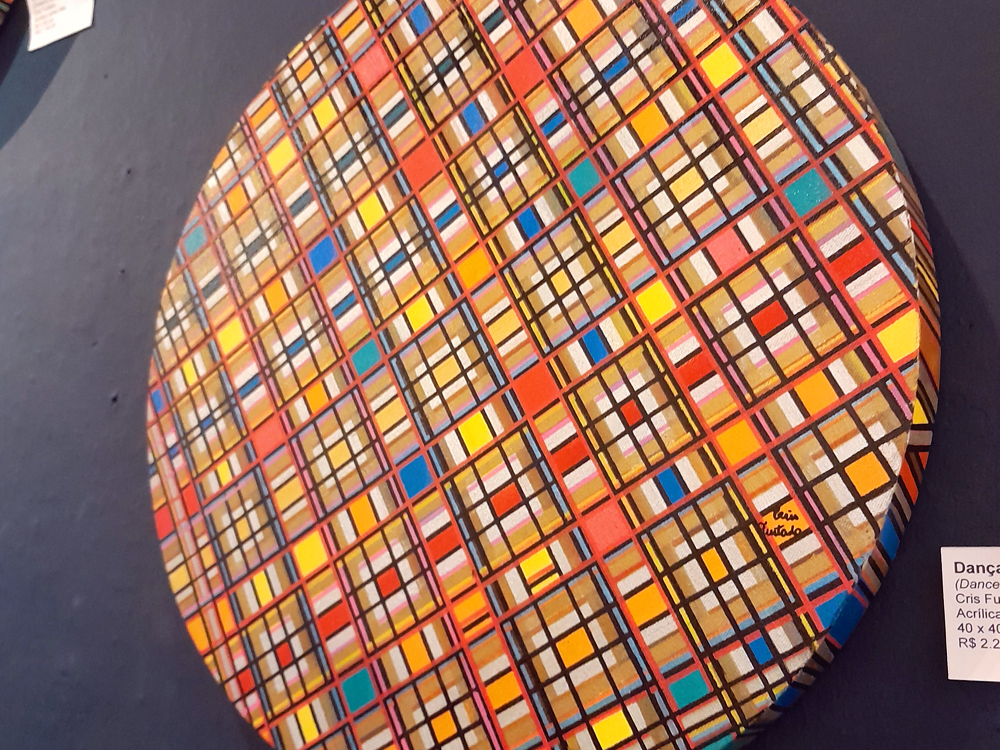 Pintura de formato circular que apresenta composição geométrica formada por linhas pretas que criam uma trama de quadrados e retângulos preenchidos por cores vibrantes, como laranja, amarelo, azul, vermelho e rosa. A obra está instalada sobre uma parede azul-escura e faz parte da exposição de artistas visuais presente na Casa Escreva, garota!