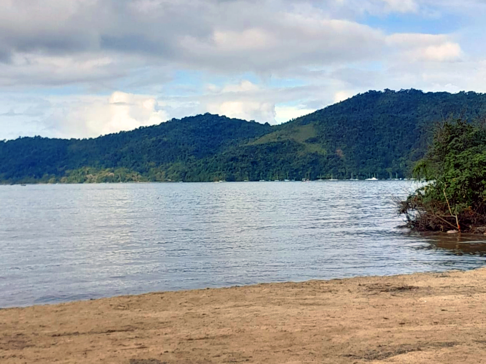 Paisagem da orla de Paraty durante a FLIP 2026. Em primeiro plano há uma faixa de areia que encontra as águas calmas da baía. Ao fundo, montanhas cobertas por Mata Atlântica se estendem até o horizonte sob um céu com nuvens claras e áreas de chuva ao longe. Pequenas embarcações aparecem próximas à costa, compondo a vista natural da cidade histórica.