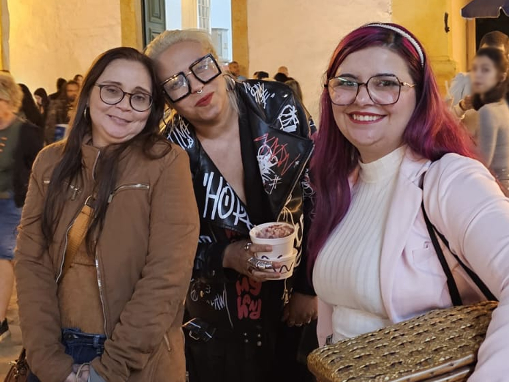 Três mulheres sorriem para a câmera durante a programação noturna da FLIP 2026, em uma rua do centro histórico de Paraty. À direita está Luly Lage, usando blazer rosa-claro sobre uma blusa branca de gola alta, cabelos longos em tom de rosa, óculos de armação grande e segurando uma caixa de palha. Ao centro, Marianna Roman veste roupa preta com detalhes em branco, usa óculos grandes de armação quadrada e segura um copo. À esquerda, sua amiga Verônica veste jaqueta marrom sobre uma blusa amarela. Ao fundo, o movimento de visitantes ocupa a rua iluminada da Festa Literária Internacional de Paraty.