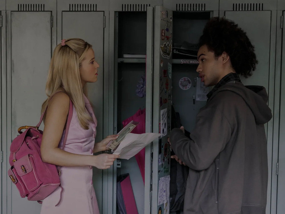 Em um corredor de escola, Elle veste um conjunto rosa-claro e carrega uma mochila rosa, conversando com um rapaz de cabelos crespos diante de armários metálicos abertos, Dustin. Ela segura alguns papéis enquanto olha para ele com expressão séria. O interior do armário ao fundo está decorado com adesivos e objetos pessoais.