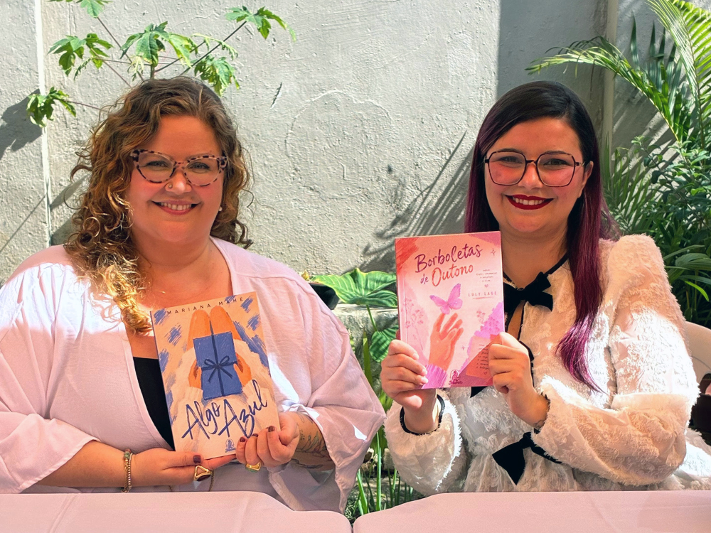 Mariana Mahé e Luly Lage posam lado a lado diante de uma mesa de autógrafos. Mariana segura seu livro Algo Azul, enquanto Luly apresenta à frente da câmera um exemplar de Borboletas de Outono, cuja capa em tons de rosa destaca uma borboleta pousando sobre uma mão. As duas sorriem para a fotografia.