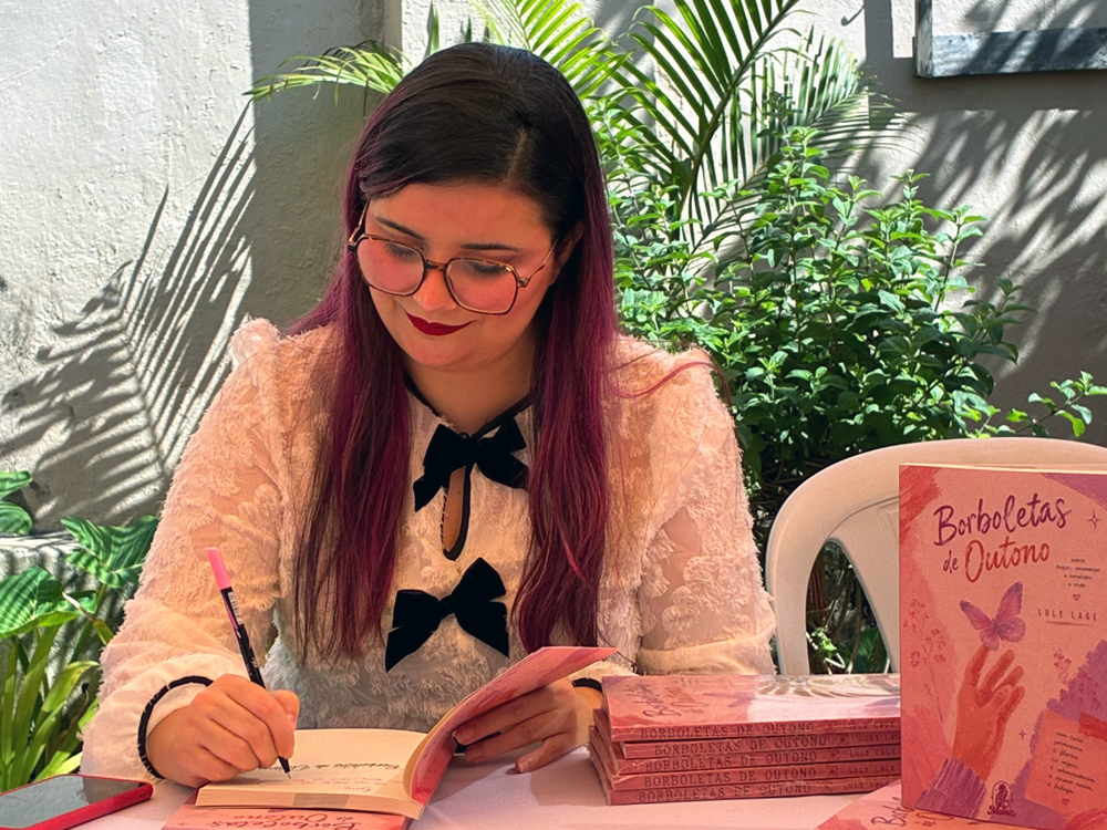 Sentada à mesa de autógrafos, Luly Lage escreve uma dedicatória em um exemplar de Borboletas de Outono usando uma caneta de tinta rosa. Ao lado, há uma pilha de livros e um exemplar em pé com a capa voltada para a câmera. A autora olha para o livro enquanto assina.