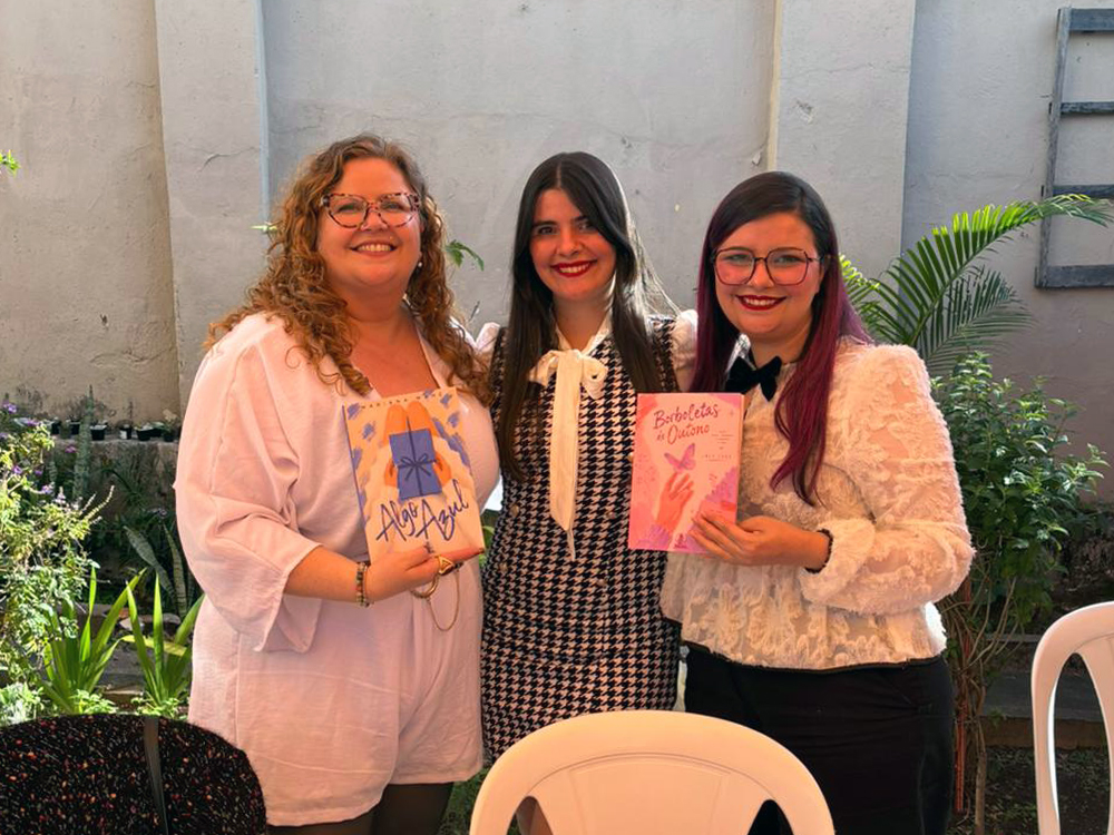 Mariana, Daninha e Luly Lage posam juntas em um jardim durante o lançamento conjunto. Mariana, à esquerda, segura um exemplar de Algo Azul. Luly, à direita, segura Borboletas de Outono. Entre elas está Daninha, irmã de Luly, sorrindo para a câmera. Ao fundo há plantas ornamentais e um muro claro, compondo um cenário ao ar livre para o evento.