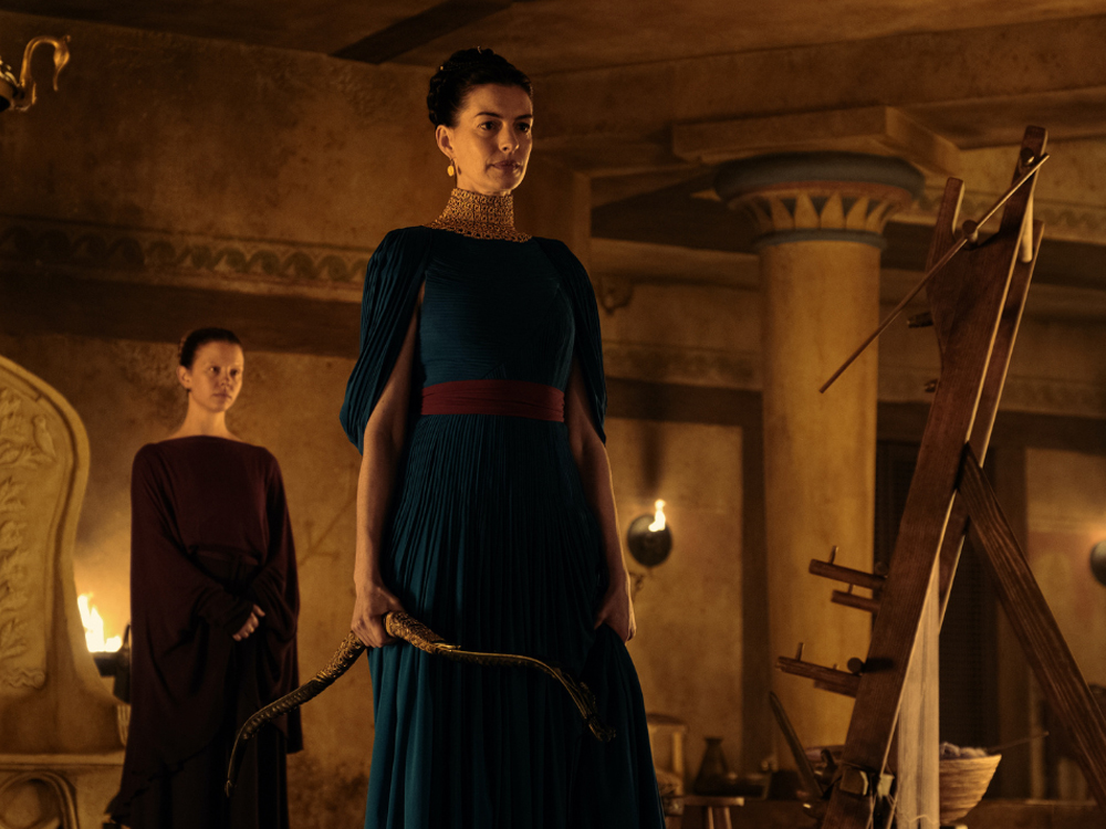 Fotografia em plano médio de Anne Hathaway interpretando Penélope em A Odisseia. Ela veste um vestido plissado azul-petróleo e segura um arco recurvo dourado e ornamentado. Atrás dela, outra mulher observa em um cenário de palácio antigo.