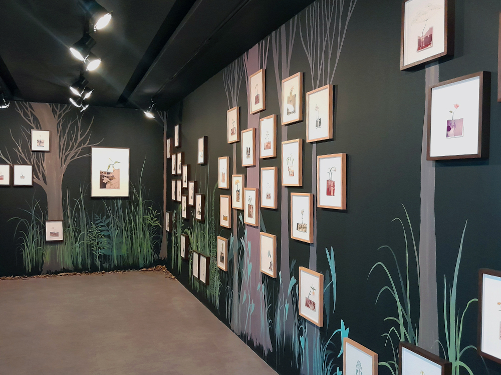 Vista geral da exposição Reflorestar, de Efe Godoy, com paredes pintadas em tons escuros e ilustrações de árvores, gramíneas e vegetação formando um cenário imersivo. Diversas obras em molduras de madeira estão distribuídas pelas paredes, iluminadas por trilhos de luz no teto.