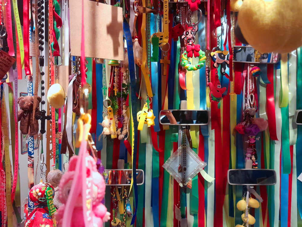 Instalação artística composta por dezenas de fitas coloridas pendentes, chaveiros, medalhas, pequenos brinquedos, amuletos e objetos do cotidiano brasileiros reunidos em uma composição vibrante e cheia de cores. Entre os elementos estão fitas religiosas, personagens da cultura pop, miniaturas e lembranças afetivas, criando um mosaico visual que remete à diversidade cultural da brasilidade.